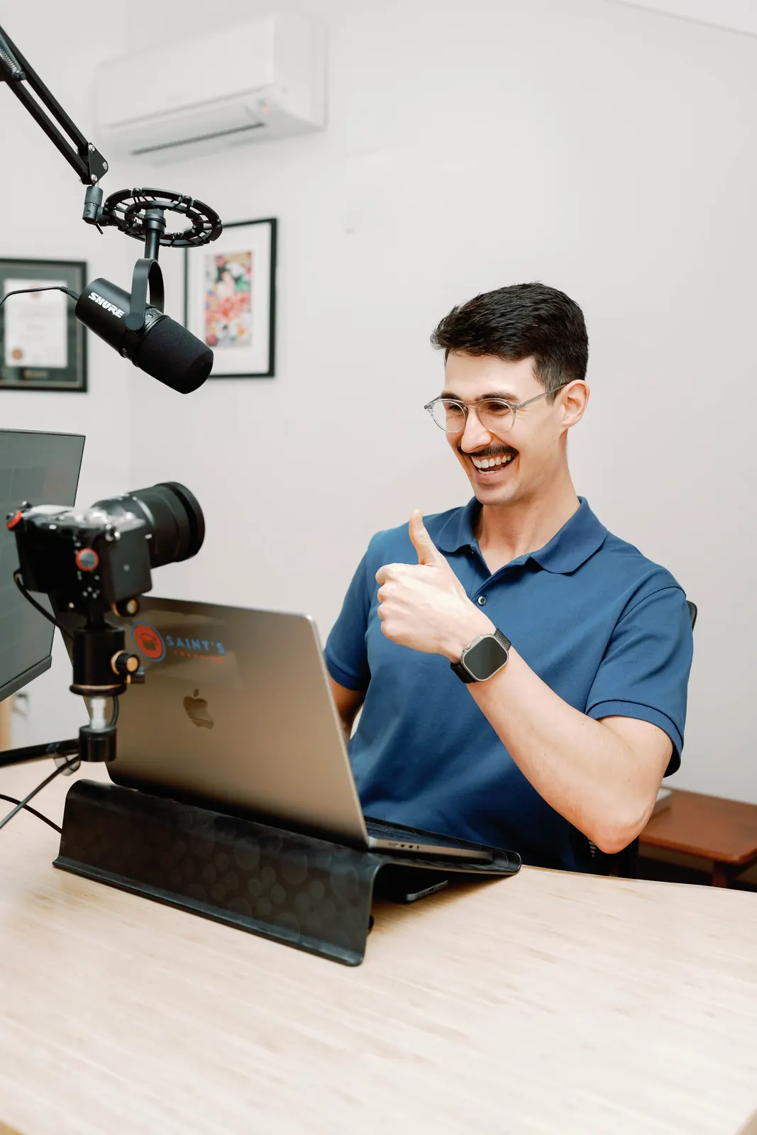 Lennox at his desk giving a thumbs up beside a laptop, a camera on a tripod and a boom-arm microphone
